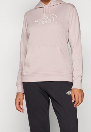 Personne portant un sweat à capuche The North Face rose clair avec une poche à l’avant et un pantalon noir avec le logo The North Face sur la cuisse.
