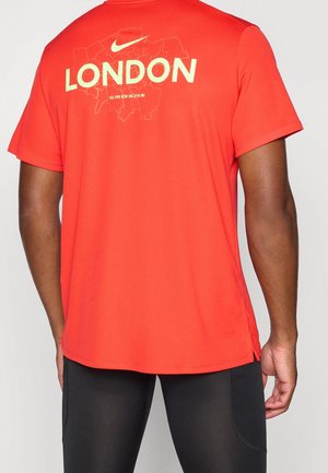 Osoba nosząca pomarańczową koszulkę Nike z "LONDON" i konturem mapy nadrukowanymi na plecach, w połączeniu z czarnymi spodniami.