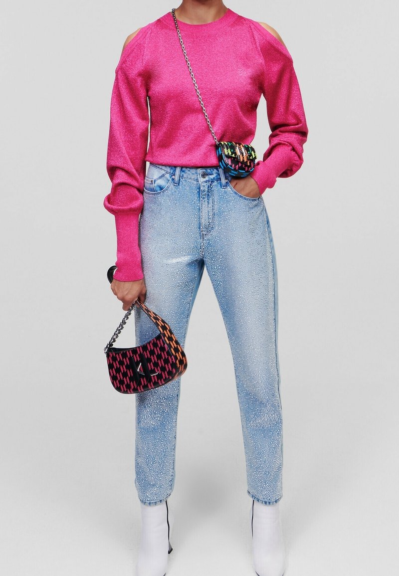 Frau trägt einen hellpinkfarbenen Cold-Shoulder-Pullover, hellblaue verzierte Jeans, weiße Stiefel und hält eine gemusterte Handtasche sowie eine kleine Umhängetasche.