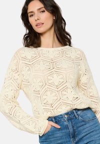 Pletena pulover v off-white barvi z odprto hexagonalno vzorcem, dolgimi rokavi in okroglim izrezom, v kombinaciji z modrimi kavbojkami.