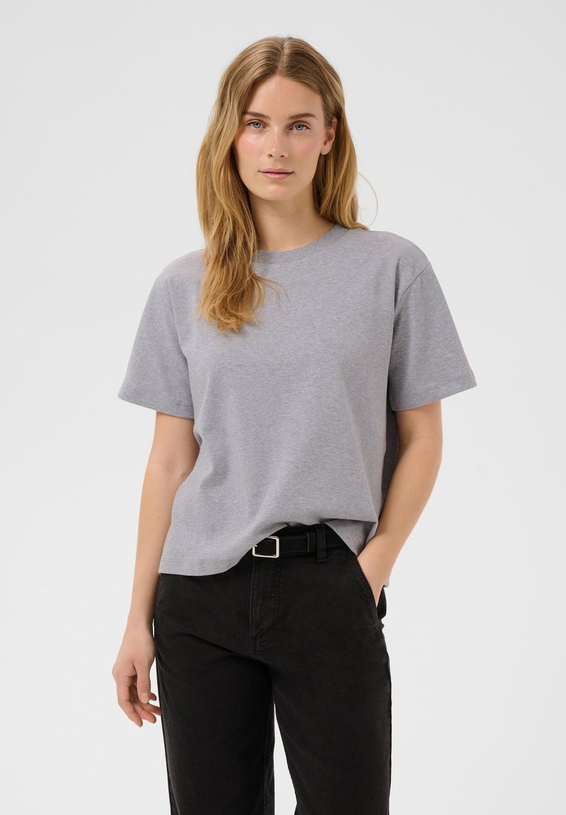 Femme aux longs cheveux blonds porte un t-shirt gris uni et un jean noir, debout avec une main dans la poche devant un fond uni.