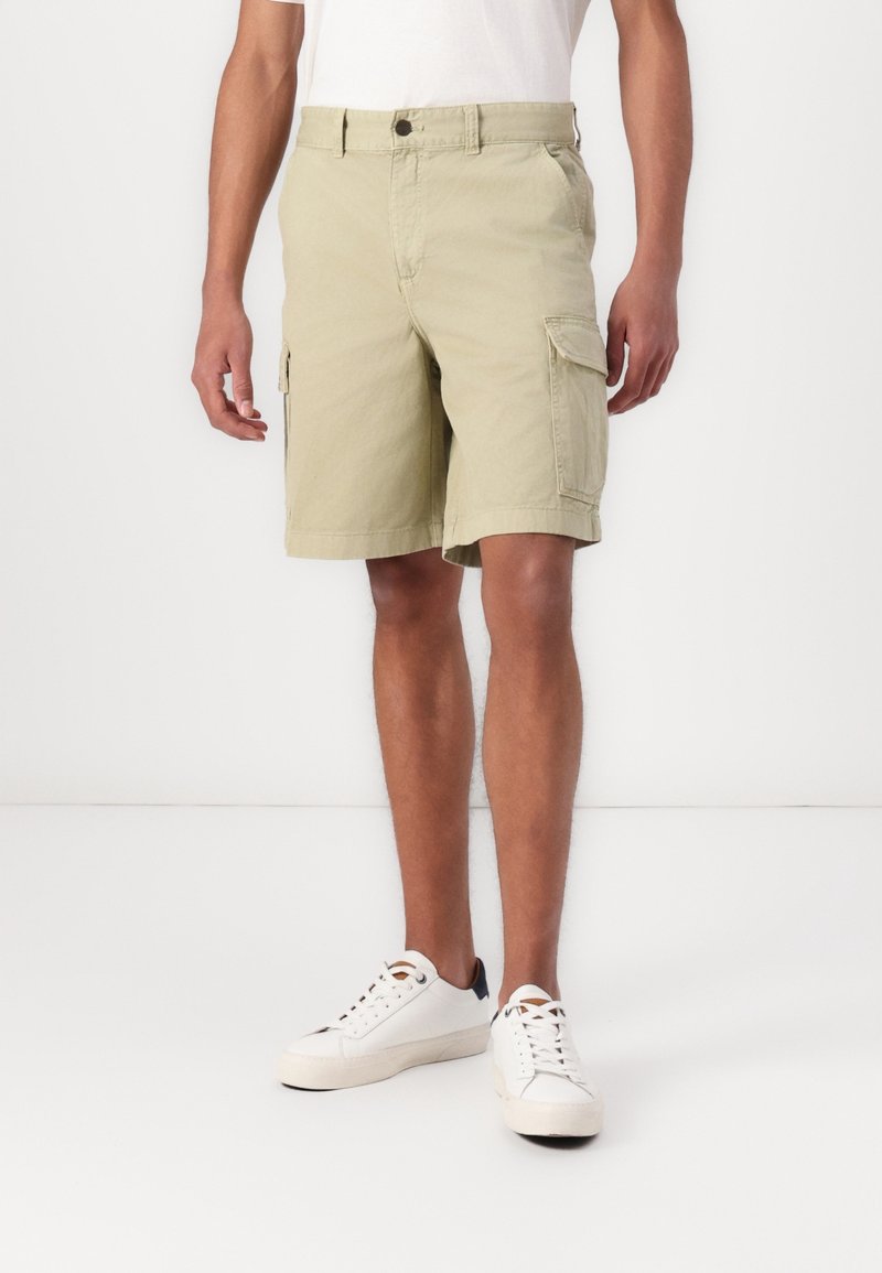 Shorts cargo vert clair longueur genou avec poches, en tissu résistant, associés à des baskets blanches. Design simple avec fermeture à bouton.