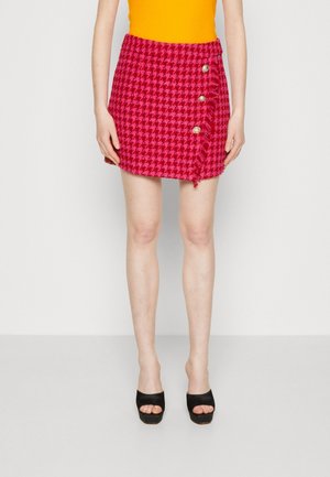 Kobieta w czerwono-różowej mini spódnicy w pepitkę z trzema złotymi guzikami z przodu oraz czarnymi sandałami na platformie z odkrytymi palcami.