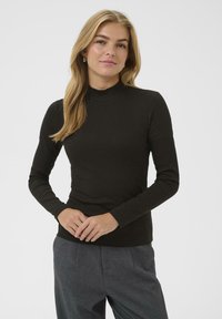 Jonge vrouw met blond haar die een zwarte lange mouwtop en grijze broek draagt, staand met de handen gevouwen voor zich tegen een effen achtergrond.