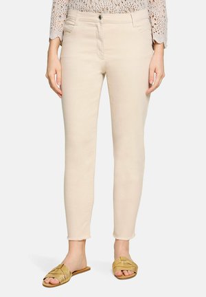 Femme portant un pantalon slim crème avec ourlet brut et des sandales dorées à enfiler, debout devant un fond clair uni.