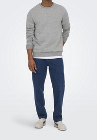 Mężczyzna w szarej bluzie z długim rękawem, niebieskich dżinsach i białych trampkach stoi na tle jasnego, gładkiego tła.