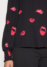 Svart långärmad skjorta med mönster av röda läppar i olika öppna och stängda uttryck, bärs med svarta byxor.