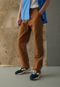 Brune bukser med en afslappet pasform, der har frontlommer og metalliske detaljer. Sættet er kombineret med mørke sneakers og en lyseblå skjorte.
