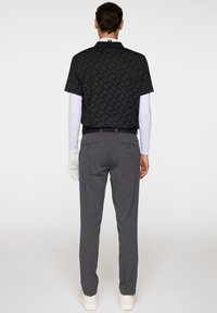 Czarna koszulka polo z krótkim rękawem w geometryczny wzór, zestawiona z szarymi spodniami. Model nosi białą drugą warstwę z długim rękawem oraz rękawice.