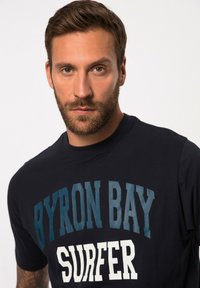 Námorně modré bavlněné tričko s krátkými rukávy, které má výrazný bílý a tyrkysový potisk s nápisem "BYRON BAY SURFER." Klasický design s kulatým výstřihem.