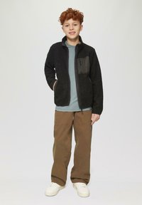 Zwarte fleece ritsjack met een gestructureerd oppervlak, grijze borstzak; gecombineerd met een lichtblauw shirt en bruine wijde broek.