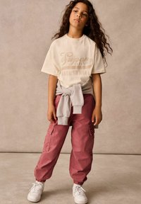 Krämfärgad t-shirt med korta ärmar och brun logotext, kombinerad med rosa cargobyxor med elastiska muddar och sidofickor, avslutad med vita sneakers.