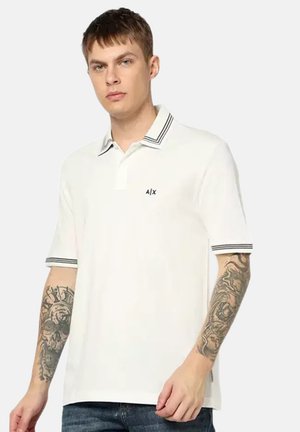 Jonge man die een wit polohemd draagt met zwarte strepen op de kraag en mouwen, met zichtbare tatoeages op beide onderarmen tegen een effen achtergrond.