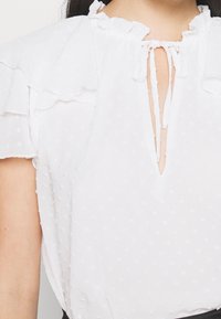 Vit texturerad blus med små upphöjda prickar, korta volangärmar och en nyckelhålsurringning som är knuten med två tunna tygsnören.