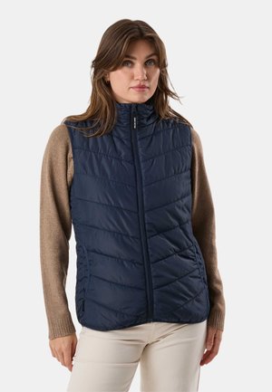 Kvinde iført en marineblå quiltet vest uden ærmer over en brun langærmet sweater og beige bukser, stående foran en ensartet lys baggrund.