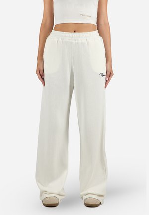 Pantalons larges blancs à côtes avec une taille élastique et un petit logo noir sur le côté gauche. Le matériau semble doux et texturé.