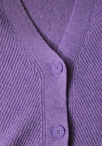 Lila kofta med V-ringning, texturerad ribbad stickning. Har två runda lila knappar framtill, slät tyg och detaljerad sömnad.