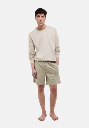 Homme debout pieds nus portant une chemise à manches longues beige et un short assorti à cordon de serrage contre un fond uni.