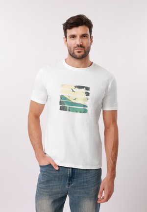 Άνδρας που φοράει λευκή μπλούζα με εικόνα βουνών, δέντρων και πουλιών σε πτήση, συνδυασμένη με μπλε τζιν, στέκεται μπροστά σε απλό φόντο.