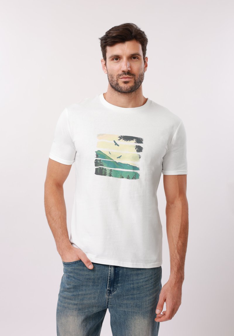 Άνδρας που φοράει λευκή μπλούζα με εικόνα βουνών, δέντρων και πουλιών σε πτήση, συνδυασμένη με μπλε τζιν, στέκεται μπροστά σε απλό φόντο.