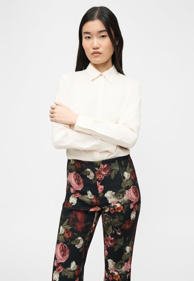 Fiatal nő hosszú sötét hajjal, krémszínű, gombos blúzt és fekete nadrágot visel piros és rózsaszín virágmintával, karjait keresztezi.