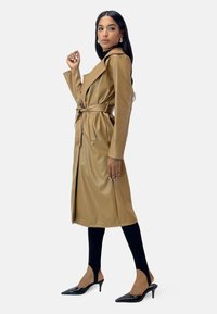 Tan faux leather trench coat with a belted waist, long sleeves, and a lapel collar. Worn over black leggings and open-toe black heels.