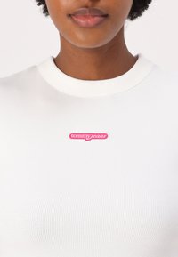 White ribbed top featuring a pink "tommy jeans" logo at the chest, round neck, and a snug fit. Soft texture with visible ribbing detail.
