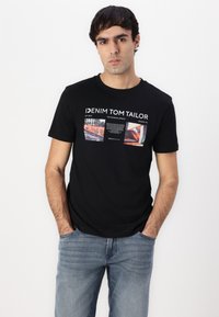 Črna bombažna majica s kratkimi rokavi, ki ima grafični potisk z barvitimi slikami in besedilom. Ima okrogli izrez, v kombinaciji z svetlo modrimi jeans hlačami.