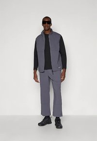 Graue Softshell-Weste über einem schwarzen Langarmshirt, kombiniert mit grauer Hose und schwarzen Sportschuhen. Glatte Textur mit Reißverschluss-Details.