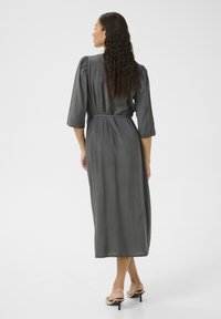 Long, gray dress with puff sleeves, a tie waist, and a flowing silhouette. Smooth fabric with a slight sheen. Worn with beige heeled sandals.