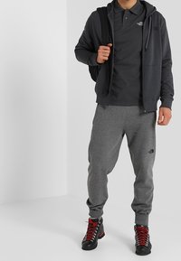 Sweat à capuche zippé gris foncé sur un polo gris, associé à un pantalon de survêtement gris et des bottes de randonnée noires avec des accents rouges. Design simple, sans motifs.