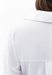 Camisa blanca con cuello y corte relajado; hecha de una tela ligera con una textura suave y detalles de costura cerca de los hombros.