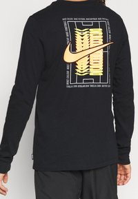 Schwarzes langärmliges Shirt mit einem großen orangefarbenen Nike-Logo und gelbem Grafikdesign auf dem Rücken, das sportliche Themen und Texturen betont.