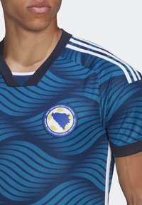 Niebieska sportowa koszulka z falistym wzorem, z okrągłym logo w kolorach niebieskim, białym i żółtym. Czarny dekolt i trzy białe paski na ramionach.