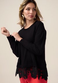 Maglione nero con polsini e bordo a coste, caratterizzato da una delicata rifinitura in pizzo floreale nella parte inferiore. Tessuto morbido, vestibilità rilassata.