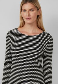 Blondynka z niebieskimi oczami, ubrana w czarną i białą, wzorzystą bluzkę z długim rękawem, stojąca na jednolitym jasnoszarym tle.
