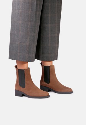 Bottines en daim marron avec panneaux latéraux élastiques noirs portées avec un pantalon large à carreaux gris sur fond blanc.