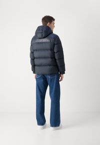 Giacca puffer blu navy con cappuccio, caratterizzata da un design trapuntato, polsini elasticizzati e una texture liscia e lucida. Abbinata a jeans a gamba larga blu.