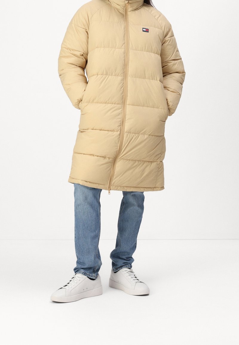 Beige polstret frakke med høj krave og lynlås, med vandrette syninger og et logo-lap på venstre bryst. Båret over blå jeans og hvide sneakers.