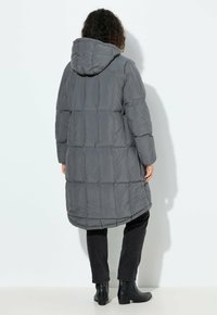 Graue Daunenjacke mit Kapuze, langer Länge und gestepptem Design. Kombiniert mit schwarzen Slim-Fit-Hosen und Ankle-Boots.
