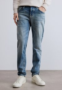 Világoskék farmernadrág halványított hatással, kopott részletekkel és egyenes szárú szabással. Fehér fűzős tornacipővel párosítva.