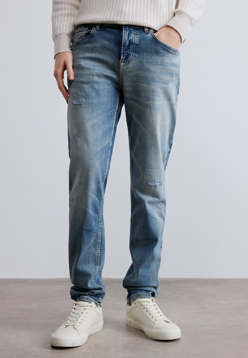 Világoskék farmernadrág halványított hatással, kopott részletekkel és egyenes szárú szabással. Fehér fűzős tornacipővel párosítva.