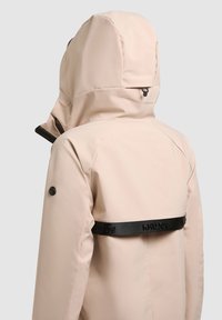 Beżowa kurtka z kapturem, widziana od tyłu, z czarnym paskiem z logo na dole pleców oraz detalem z guzikiem na lewym rękawie.