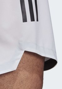 Pantaloni corti sportivi bianchi realizzati in tessuto testurizzato, con tre strisce verticali nere e un orlo in contrasto con una leggera curvatura sul bordo.