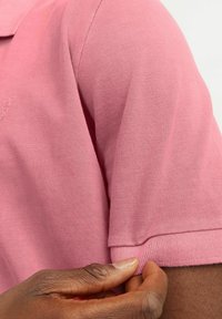 Polo majica s kratkimi rokavi v mehki roza tkanini, ki ima teksturirano pletenje, rebrast ovratnik in izvezeno logo na prsih.