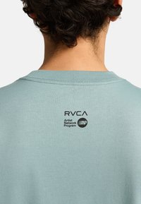 T-shirt azzurro chiaro con collo a giro, realizzata in cotone. Presenta un logo e del testo stampati in nero sul retro in alto, con una texture liscia.