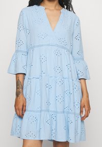 Ljusblå klänning med V-ringning, trekvarts flareärmar, broderade ögonlapp detaljer och lager på lager volanger längst ner.