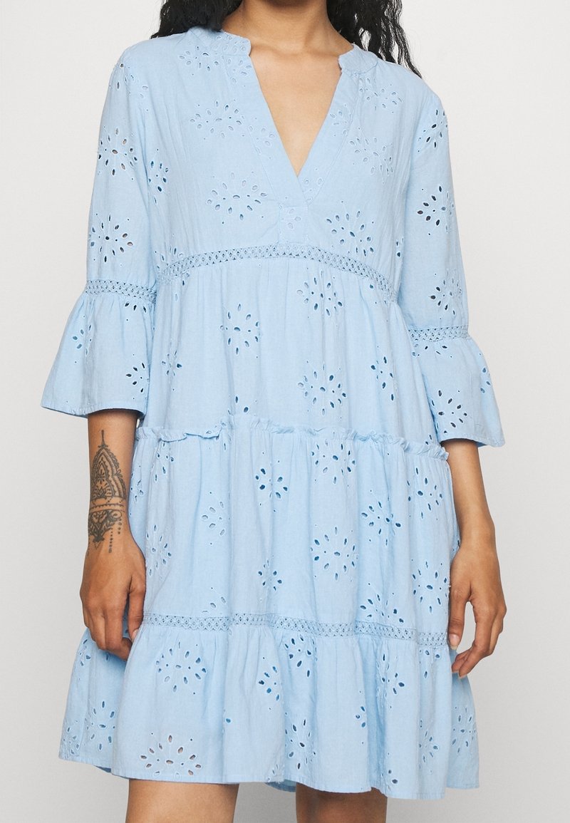 Ljusblå klänning med V-ringning, trekvarts flareärmar, broderade ögonlapp detaljer och lager på lager volanger längst ner.