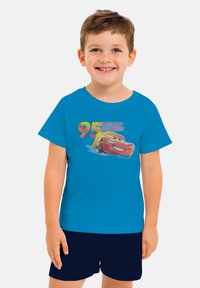 Ragazzo giovane sorridente che indossa una maglietta blu con una grafica colorata della macchina Lightning McQueen e il numero 95, abbinata a pantaloni corti scuri.