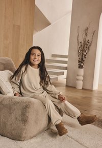 Completo beige a coste di abbigliamento comodo per bambini, con maniche lunghe e pantaloni. La bambina è seduta su un divano soffice color taupe, indossando stivali marroni.